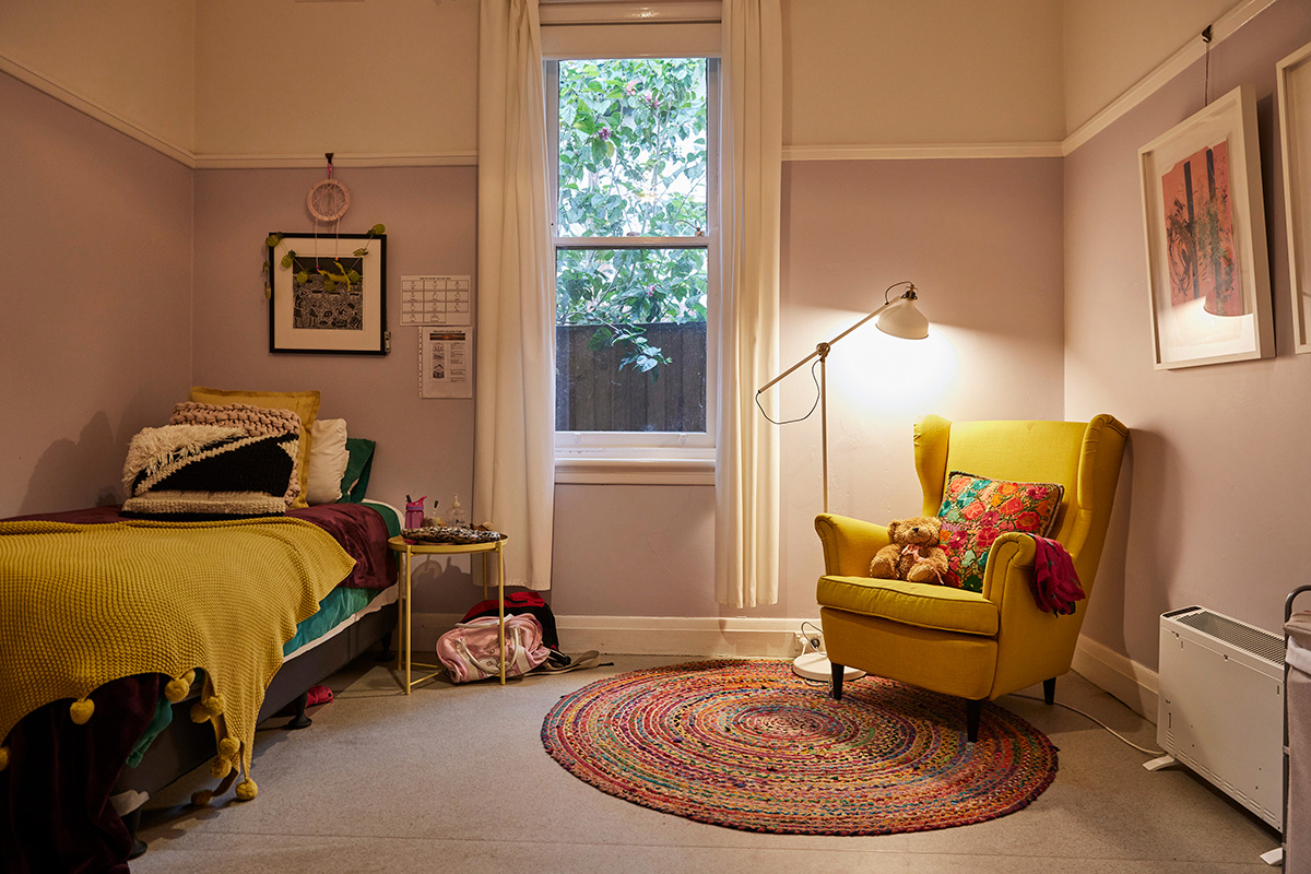 Image is of a bedroom in Rosevilla house. There is a single bed to the left of the image and a single chair to the right with a light above